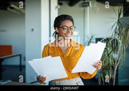 Erfolgreiches Unternehmertum. Glückliche afroamerikanische Geschäftsfrau, die im Büro Finanzpapiere überprüft Stockfoto