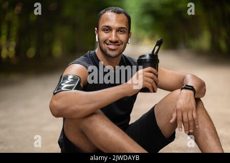 Entspannter afroamerikanischer Sportler, der auf dem Boden sitzt und Protein trinkt Stockfoto