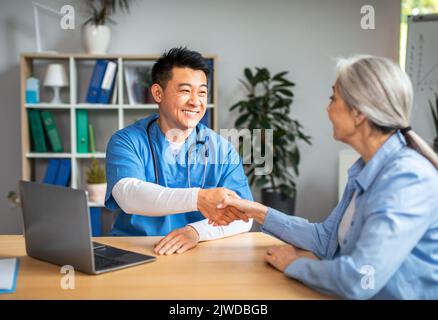 Lächelnder reifer chinesischer Mann Arzt schüttelt sich die Hände mit einer älteren kaukasischen Frau in der Klinik Stockfoto