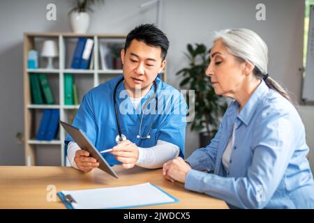 Ernst mittleren Alters asiatische Mann Arzt zeigt Tablette zu pensionierten europäischen Frau Patienten in der Klinik Stockfoto