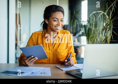 Positive schwarze Frau, die ein digitales Tablet und einen Laptop verwendet, im Büro online arbeitet und mit Kollegen per Videoanruf abruft Stockfoto