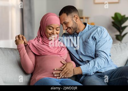 Verheiratetes schwarzes muslimisches Paar, das Baby erwartet, das im Wohnzimmer auf der Couch kuschelt Stockfoto