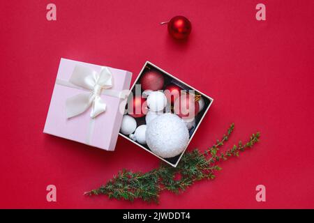 Draufsicht auf Weihnachtsdekorationen in offener Geschenkbox und grünem Christbaumzweig auf rotem Hintergrund. Schöne weiße runde Spielzeuge in rosa Verpackung. Merr Stockfoto