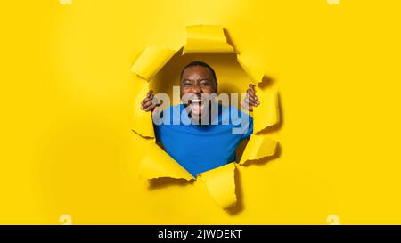 afroamerikanischer Mann, der schrie, reagierte auf etwas erstaunliches, das durch ein Loch im gelben Papierhintergrund posierte, Panorama Stockfoto