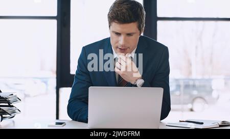 Nachdenklicher Geschäftsmann, der das Kinn berührt und den Laptop im Büro anschaut, Bild aus dem Stock Stockfoto