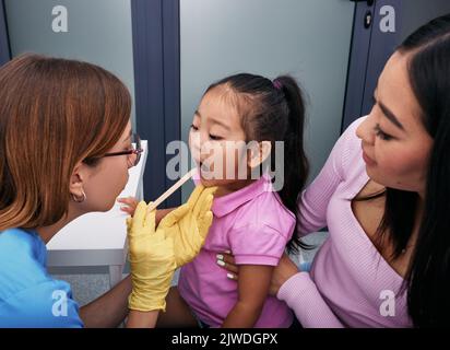 Der Arzt benutzt einen Inspektionsspachtel, um die Kehle eines koreanischen Mädchens zu untersuchen. Niedliches weibliches Kind mit ihrer Mutter beim Kinderarzt-Termin Stockfoto