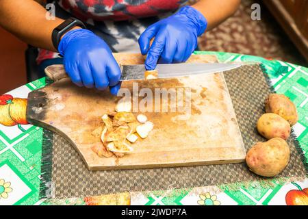 Eine nicht erkennbare Lateinerin schälte Kartoffeln mit blauen Handschuhen in ihrer Küche zu Hause Stockfoto