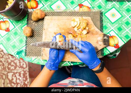 Draufsicht auf eine nicht erkennbare lateinerin, die Kartoffeln schält und blaue Handschuhe trägt Stockfoto