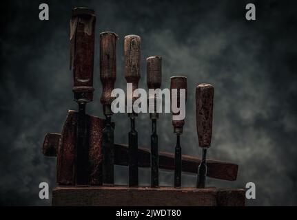 Set aus Schnitzwerkzeugen mit alten rostigen Zischen und Hammer hinter der hölzernen Werkbank vor dunklem Texturhintergrund Stockfoto