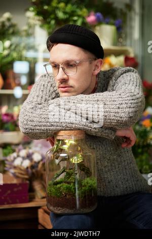 Kaukasische müde Hipster Unternehmer Floristen männlich trägt Pullover, Mütze und Brille im Blumenladen. Schöner Gewächshausbesitzer oder Gärtner mit Pflanzen in einem Glas. Hochwertiges vertikales Bild Stockfoto