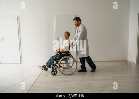 Professioneller männlicher Arzt, der weibliche Patientin auf dem Rollstuhl in der Medizinklinikhalle trägt Stockfoto