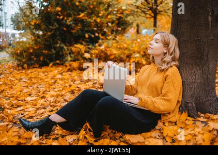 Ein schönes Mädchen Teenager sitzt in einem Herbstpark mit einem Laptop. Der Student studiert auf der Straße. Das Mädchen schloss die Augen und ruhte Stockfoto