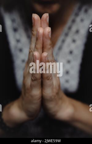 Nahaufnahme einer Frau, die mit ihren Händen zusammen betet oder meditiert, in namastiger Geste und im Hintergrund verschwommener Körper in einer dunklen launischen Atmosphäre Stockfoto
