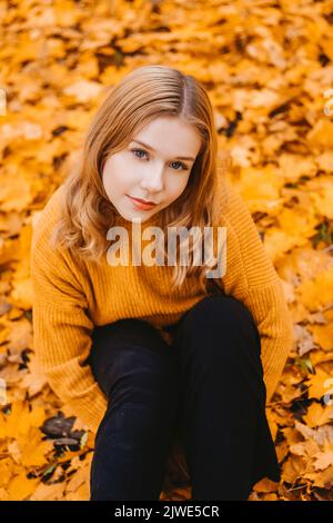 Ein Mädchen in einem weißen Pullover sitzt auf den Herbstblättern. Eine schöne Blondine mit einem Quadrat schaut auf die Kamera. Frau im Freien im Herbstpark. Stockfoto