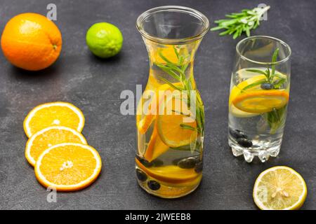 Orange, Heidelbeeren und Rosmarin infundiert Wasser in Glas und Flasche. Orange und Limette auf dem Tisch. Schwarzer Hintergrund Stockfoto