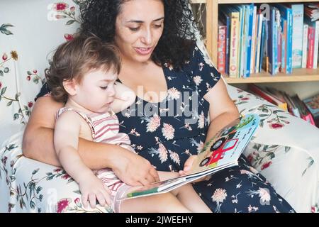 Eine junge Mutter will ihr kleiner Kleinkind-Sohn saß auf ihrem Schoß, als sie ein Bilderbuch liest Stockfoto