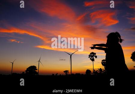 Silhouette von zwei Mädchen, die bei Sonnenuntergang auf Windturbinen zeigen, Jeneponto, Süd-Sulawesi, Indonesien Stockfoto