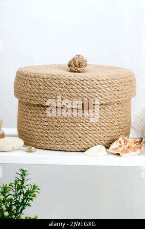 Handgestrickter Jute-Korb mit Deckel im Badezimmer auf einem weißen Holzregal. Stockfoto