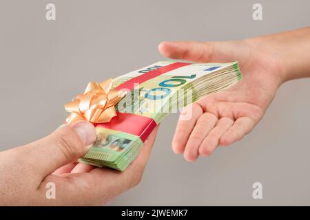 Geld als Geschenk, Preis oder Bonus. Ein Mann gibt einem Kind ein Geldbündel. Banknoten von 100 Euro sind mit einem roten Band mit einem Bogen gebunden. Stockfoto