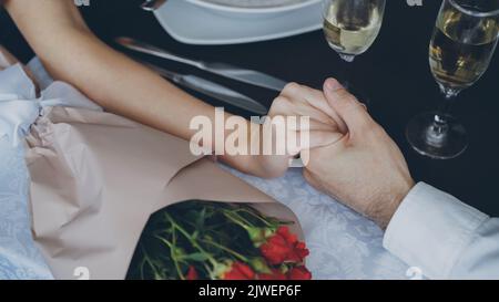 Nahaufnahme der männlichen Hand, die die gepflegte weibliche Hand mit Sektgläsern und Blumen auf dem Tisch hält und zusammendrückt. Romantische Beziehung, Liebe und feines Dining-Konzept. Stockfoto