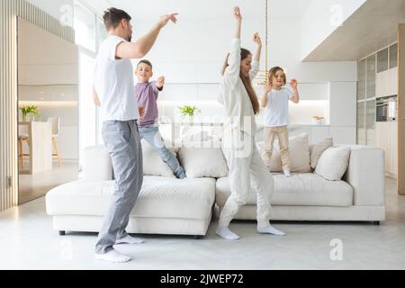 Lustige aktive Familie aus vier jungen erwachsenen Eltern und niedlichen kleinen Kindern, die im Wohnzimmer zusammen tanzen, sorglose kleine Kinder mit Mama Papa ha Stockfoto