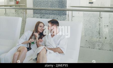 Das junge glückliche Ehepaar sitzt entspannt im modernen Spa-Salon mit Cocktailgläsern und plaudert. Romantische Beziehung, Entspannung und Wellness-Konzept. Stockfoto