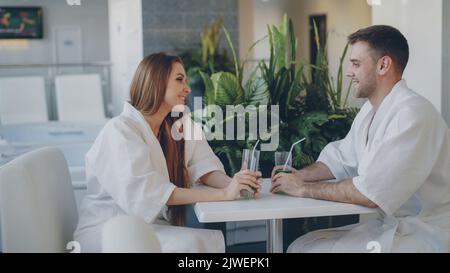 Fröhliche junge Leute reden und trinken, während sie im Day Spa am Tisch sitzen. Komfortable weiße Möbel und grüne Pflanzen sind sichtbar. Stockfoto