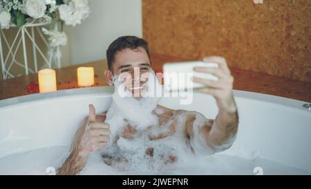 Gutaussehender, fröhlicher Kerl nimmt Selfie in der Badewanne mit Seifenschaum auf seinem Bart mit einem Smartphone auf. Er lacht und macht Gesten mit seiner Hand, die posiert und Spaß hat. Stockfoto