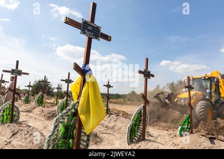 Ein Grabkreuz, das mit einer ukrainischen Flagge gebunden ist, vor dem Hintergrund eines Baggers, der Gruben für frische Gräber gräbt. Begräbnis der Überreste von 13 nicht identifizierten und zwei identifizierten Personen, die während der russischen Besatzung am 2. September 2022 im Bezirk Buchan getötet wurden. Wenige Monate nach der Entbesetzung des Bezirks Bucha wurden die nicht identifizierten Leichen auf dem Friedhof in Bucha begraben und mit Zahlen gekennzeichnet. Nach Angaben des stellvertretenden Bürgermeisters von Buch Mykhailyna Skoryk wurden insgesamt 419 Menschen, die während der russischen Besetzung des Bezirks Buchan getötet wurden, in Th begraben Stockfoto