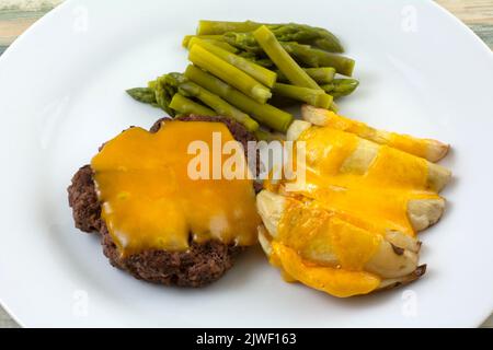 Warme Mahlzeit aus Hamburger-Patty und Kartoffelkeilen mit geschmolzenem Cheddar-Käse und Spargel auf weißem Teller Stockfoto