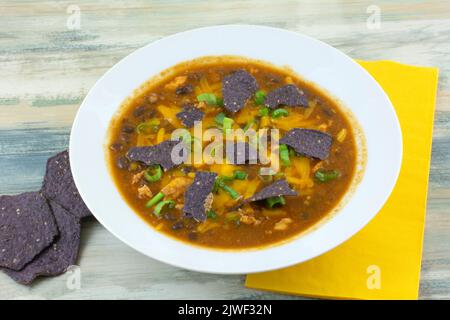 Hühnchen- und Schwarzbohnensuppe mit blauen Mais-Tortilla-Chips in weißer Schüssel mit gelber Serviette Stockfoto