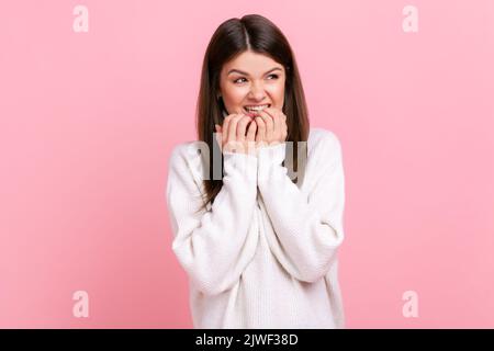 Hübsche Brünette Frau beißt Nägel, nervös erschrocken, Gefühl Angst, Angststörung, trägt weißen lässigen Stil Pullover. Innenaufnahme des Studios isoliert auf rosa Hintergrund. Stockfoto