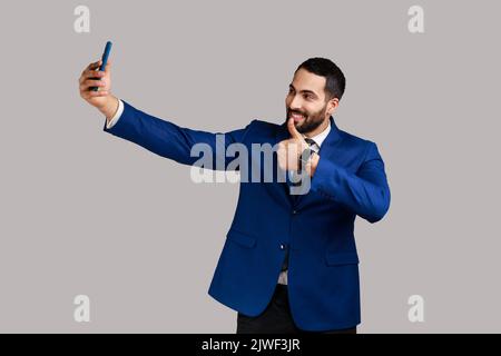 Positiver Mann, der Selfie auf der Smartphone-Kamera macht, Blogger kommuniziert, Video für Follower aufzeichnet, Daumen hochzeigt, offiziellen Style-Anzug trägt. Innenaufnahme des Studios isoliert auf grauem Hintergrund. Stockfoto