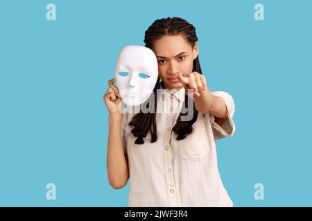 Streng herrisch Frau mit schwarzen Dreadlocks hält weiße Maske in der Hand, zeigt den Finger auf die Kamera mit ernstem Ausdruck, trägt weißes Hemd. Innenaufnahme des Studios isoliert auf blauem Hintergrund. Stockfoto