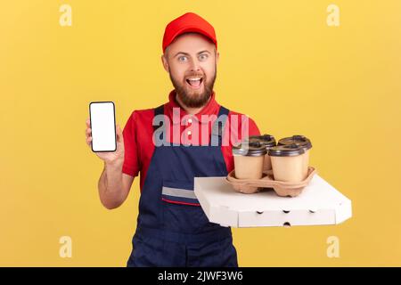 Porträt von staunenden aufgeregt Kurier Mann mit Pizza-Box und Kaffee in Einweg-Tasse stehen und zeigt Telefon mit leerem Bildschirm für Werbung. Innenaufnahme des Studios isoliert auf gelbem Hintergrund Stockfoto