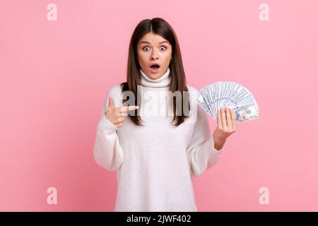 Schockiert lächelnde Frau zeigt auf Fan von Dollar-Banknoten, schaut überrascht auf die Kamera, trägt weißen Pullover im lässigen Stil. Innenaufnahme des Studios isoliert auf rosa Hintergrund. Stockfoto