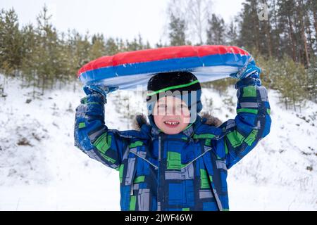 Fröhlicher, aktiver Junge, der draußen einen aufblasbaren Schlauch über den Kopf hält. Nettes kleines glückliches Kind, das im Winter Spaß im Freien auf Schlitten hat. Familie Stockfoto