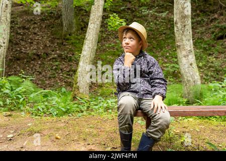 Lustiger kaukasischer Junge in einem Hut des Reisenden und Windbreaker sitzt auf einer Bank Stockfoto