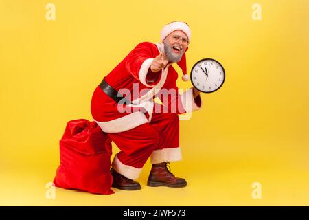 Fröhlicher, positiver älterer Mann mit grauem Bart im weihnachtsmann Kostüm sitzt auf einer roten Tasche mit Geschenken, hält die Wanduhr in den Händen und zeigt auf die Kamera. Innenaufnahme des Studios isoliert auf gelbem Hintergrund Stockfoto