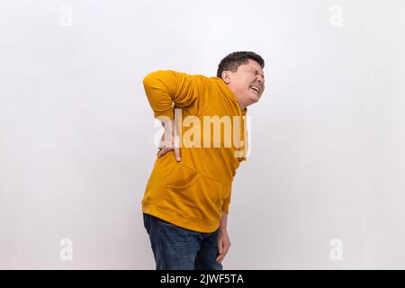 Wirbelsäulenprobleme. Mann mittleren Alters, der seinen Rücken hält, plötzliche Schmerzen im unteren Rücken, eingeklemmte Nerven- oder Nierenentzündungen hat und einen Kapuzenpullover im urbanen Stil trägt. Innenaufnahme des Studios isoliert auf weißem Hintergrund. Stockfoto