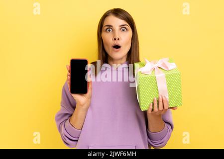 Present, Bonus für mobile Benutzer. Porträt einer verblüfften, überraschten Frau, die eine Geschenkbox und ein Mobiltelefon mit einem Mock-up, leerem Display und einem Hoodie hält. Innenaufnahme des Studios isoliert auf gelbem Hintergrund. Stockfoto