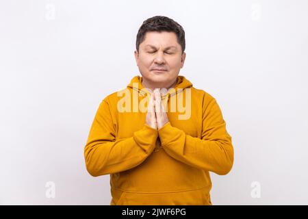 Yoga-Zeit. Porträt eines gutaussehenden, ruhigen Mannes mittleren Alters, der in Yoga-Pose steht und versucht, sich zu entspannen, hält die Handflächen zusammen und trägt einen Hoodie im urbanen Stil. Innenaufnahme des Studios isoliert auf weißem Hintergrund. Stockfoto