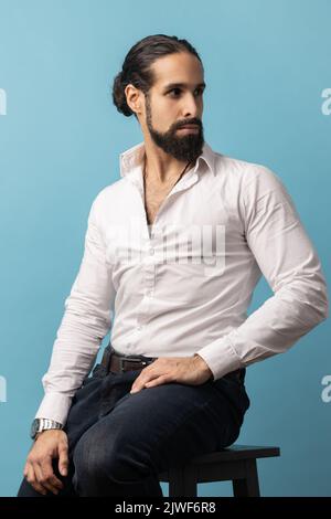 Porträt eines attraktiven Mannes mit Bart und dunklen Haaren in weißem Hemd und schwarzer Hose, sitzend und mit selbstbewusstem Ausdruck wegblickend. Innenaufnahme des Studios isoliert auf blauem Hintergrund Stockfoto