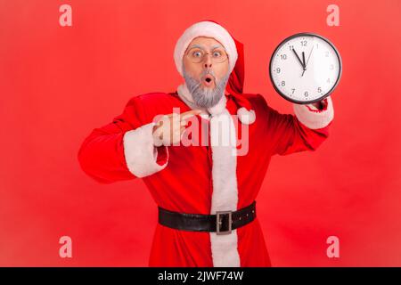 Porträt eines schockierten älteren Mannes mit grauem Bart, der das weihnachtsmann-Kostüm trägt und auf die Wanduhr zeigt, die auf einen Termin wartet und den Mund offen hält. Innenaufnahme des Studios isoliert auf rotem Hintergrund. Stockfoto