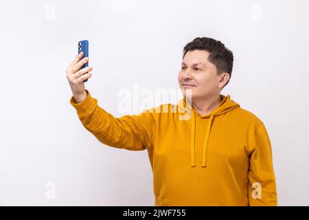 Porträt eines fröhlichen Mannes, der mit dem Smartphone Selfie macht, bei einem Videoanruf spricht und fröhlich lächelt, mobile Kommunikation, mit einem Hoodie im urbanen Stil. Innenaufnahme des Studios isoliert auf weißem Hintergrund. Stockfoto