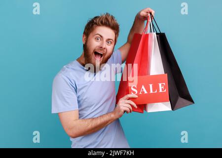 Porträt eines gutaussehenden bärtigen Mannes, der Papiertaschen und Verkaufsschild in der Hand hält, Einkaufen und Rabatte, und mit aufgeregt Ausdruck auf die Kamera blickt. Innenaufnahme des Studios isoliert auf blauem Hintergrund. Stockfoto