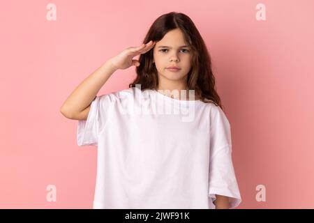 Porträt eines ernsthaften kleinen Mädchens, das weißes T-Shirt trägt und einen Gruß gibt, gehorsam dem Befehl des Befehlshabers mit einem aufmerksamen, selbstbewussten Gesicht zuhört. Innenaufnahme des Studios isoliert auf rosa Hintergrund. Stockfoto