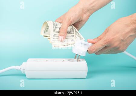 Frau hält einen Netzstecker und US-Dollar-Banknoten in der Nähe des Verlängerungskabels auf blauem Hintergrund. Stromkosten, Strompreise Stockfoto