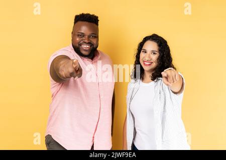 Porträt eines lächelnden, zufriedenen Paares in legerer Kleidung, das zusammen steht und mit Zeigefinger auf die Kamera zeigt und positive Emotionen ausdrückt. Innenaufnahme des Studios isoliert auf gelbem Hintergrund. Stockfoto