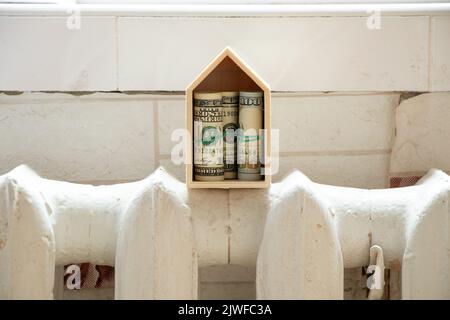 Ein kleines Holzhaus mit hundert Dollar Scheinen im Inneren steht auf einer gusseisernen Batterie in der Wohnung, Heizperiode, eine warme Batterie in der Wohnung Stockfoto